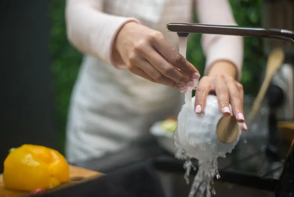 Close-up of hands washing a white bowl under a running faucet in a kitchen.